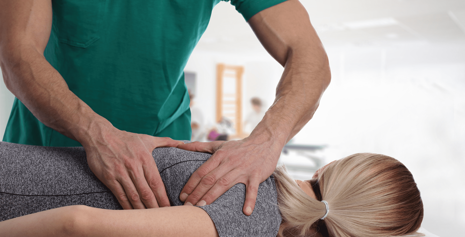 A person receives a back adjustment from a practitioner in a green uniform, in a well-lit clinic setting.
