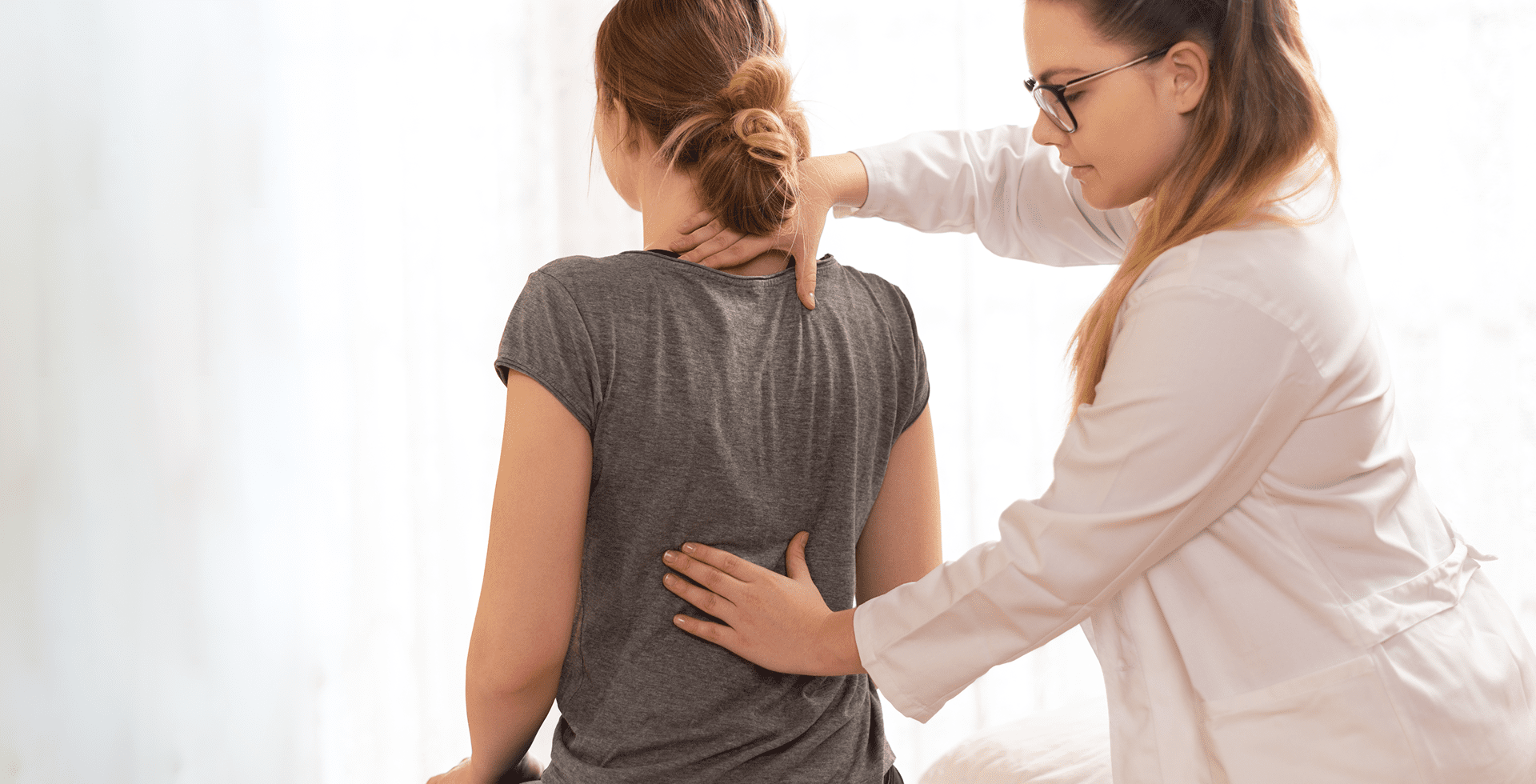 A healthcare professional examines a seated woman's back, placing one hand on her neck and the other on her lower back, in a light-filled room.