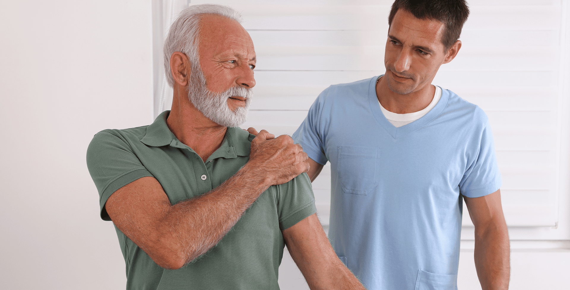 An older man in a green shirt holds his shoulder, while a younger man in a blue shirt looks at him, offering assistance in a bright room.