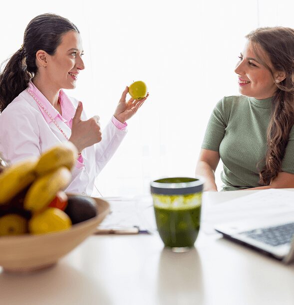 Rectangle 29 Doctor discussing nutrition with a patient, holding a lemon and giving a thumbs up. A bowl of fruit and a green smoothie are on the table in the foreground.