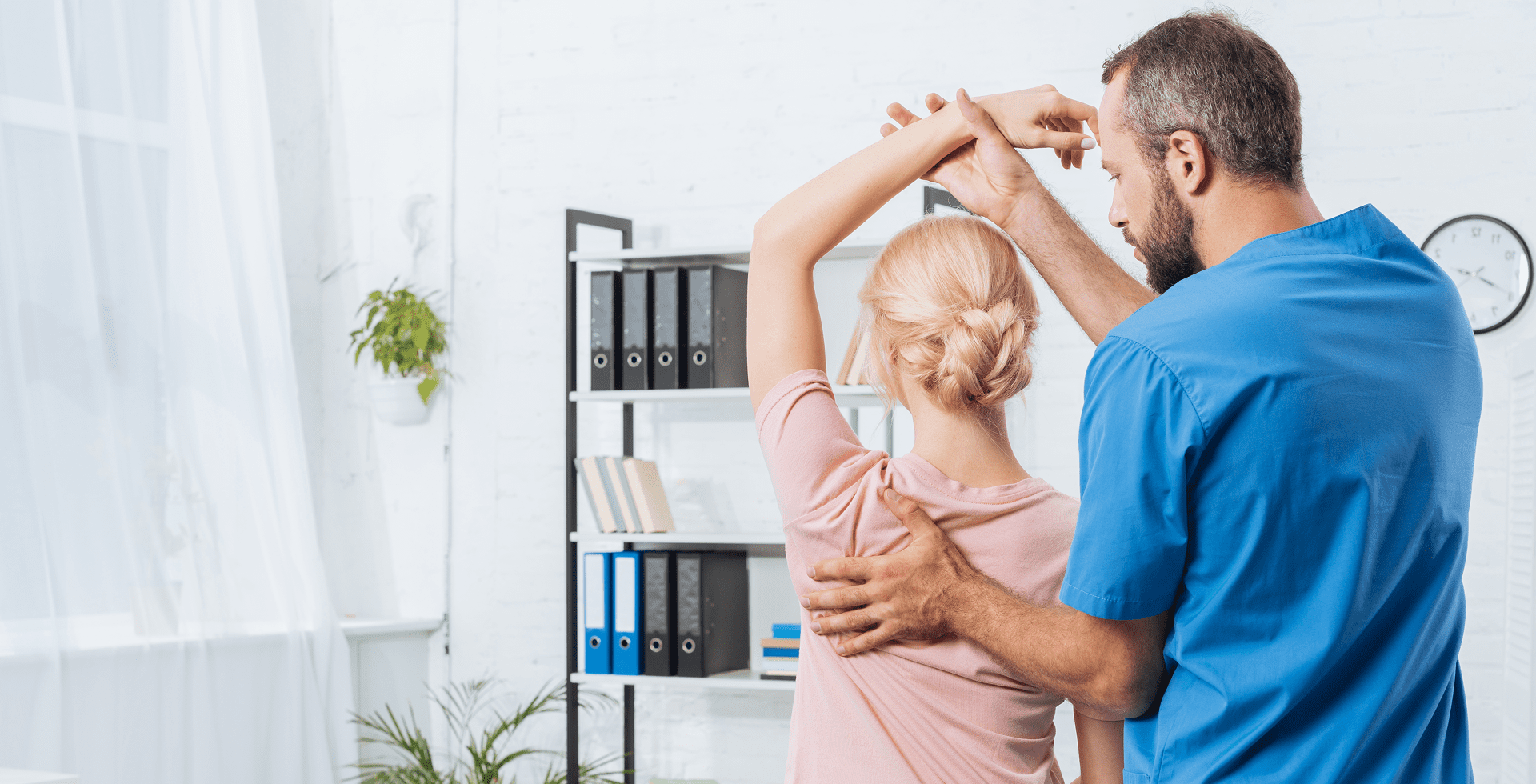A physiotherapist assists a patient with arm stretching exercises in a bright room with shelves and plants.